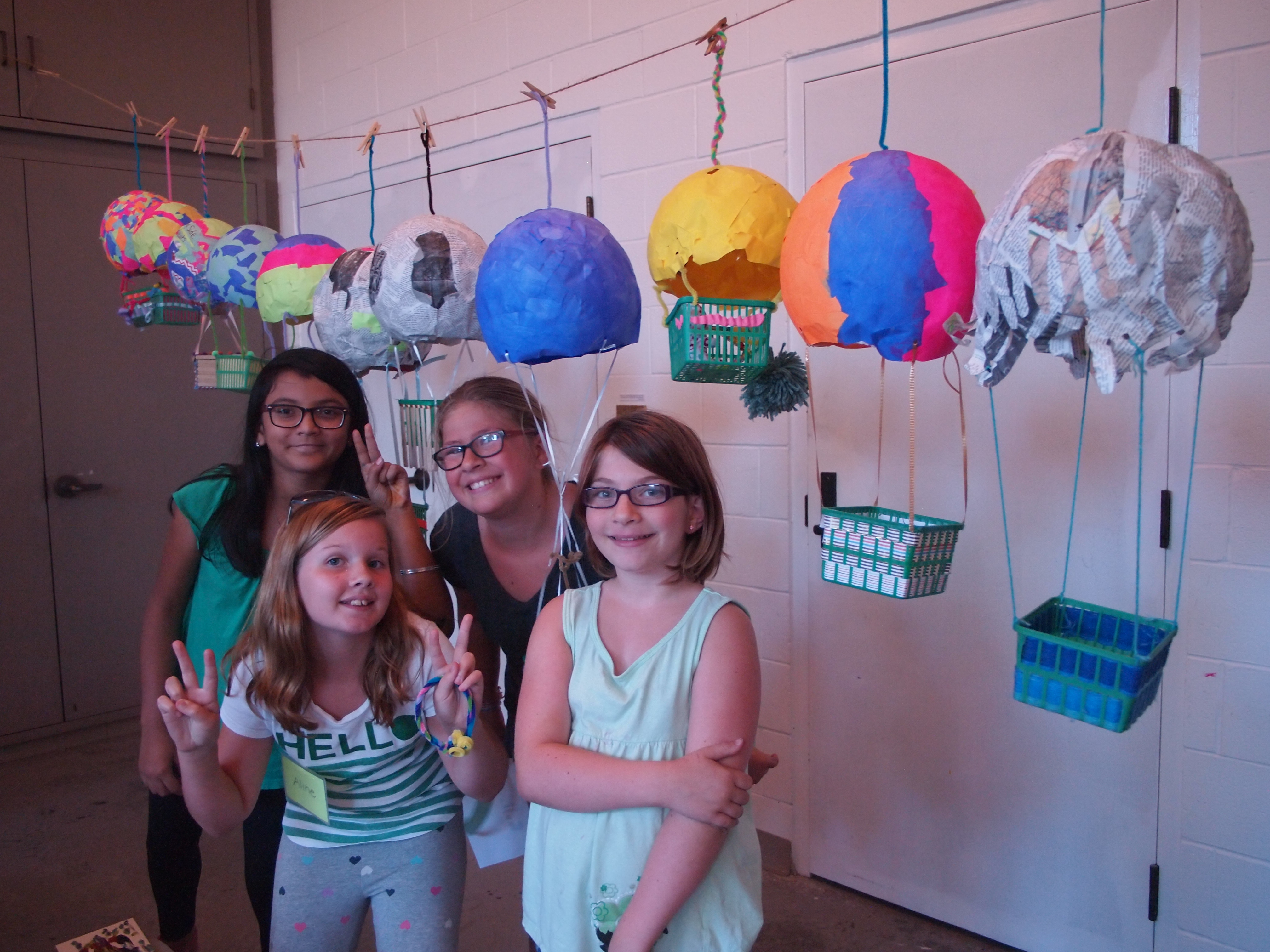 A group of four girls posing beneath a string hung with various brightly coloured hot air balloons made from paper and produce baskets