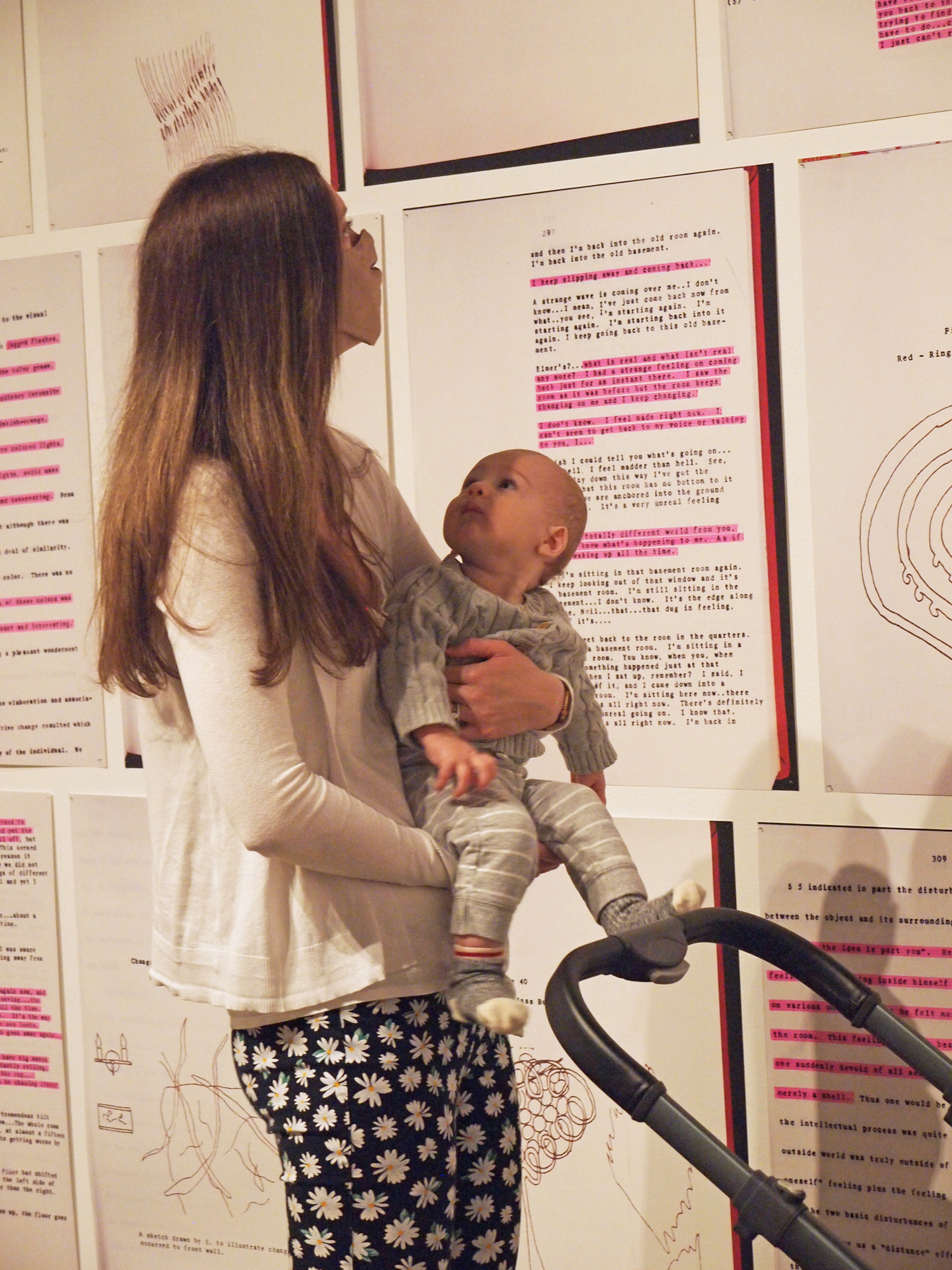 A mother holds her baby while looking at a wall filled with large text prints with pink highlighted segments