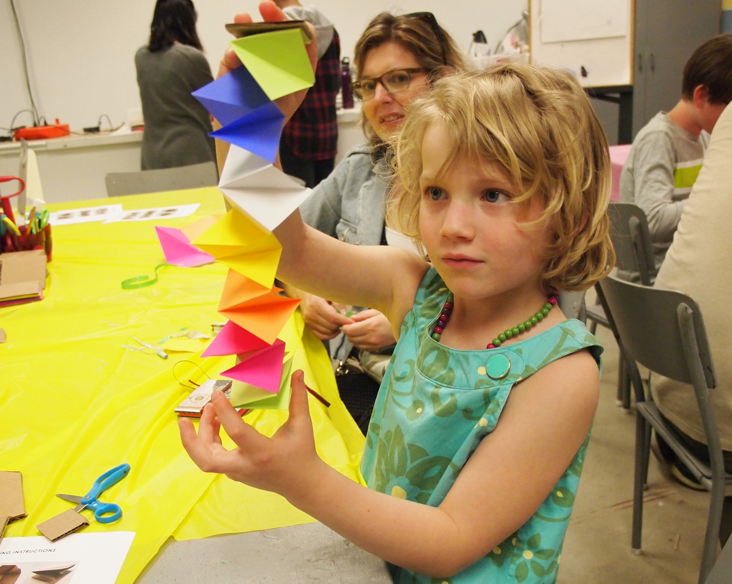 A young girl extends a colourful folded paper sculpture vertically between her hands while her mother looks on in the KWAG studio