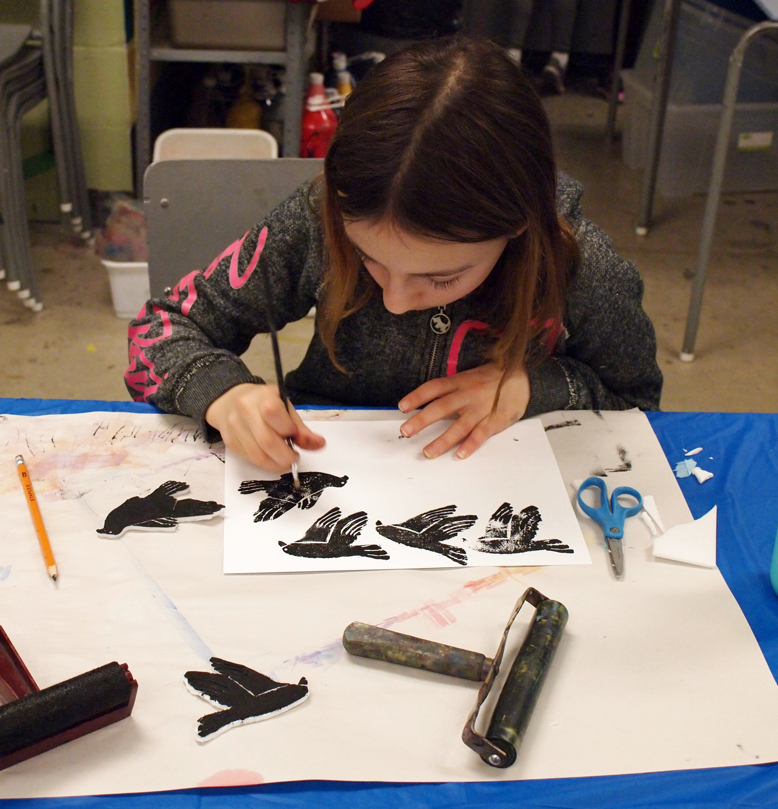 A young girl uses a brush to touch up a print in progress of repeating black bird patterns with printmaking tools laid out around her work space