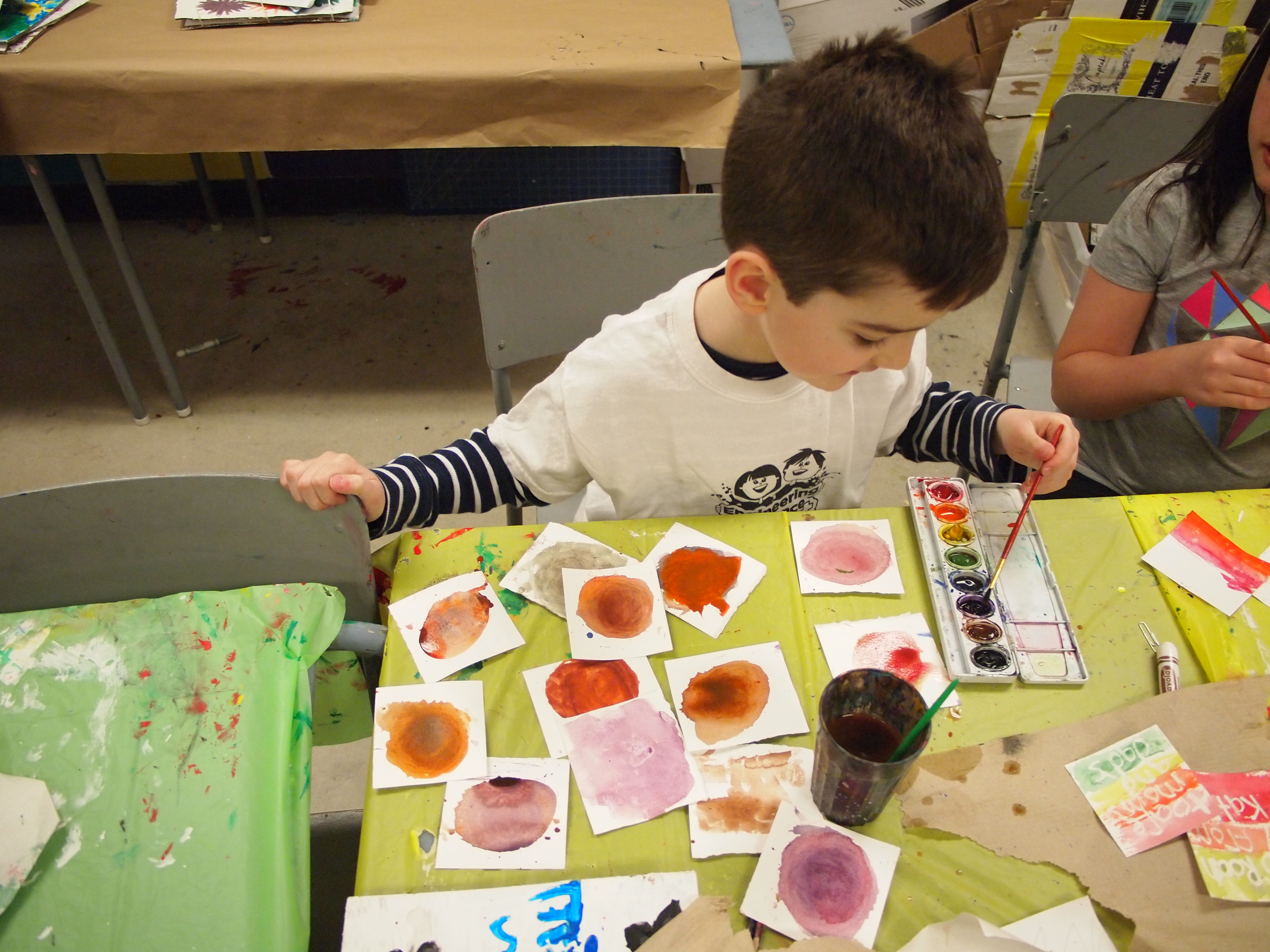 A young boy experiments with painting many coloured swatches on small pieces of paper in the KWAG studio