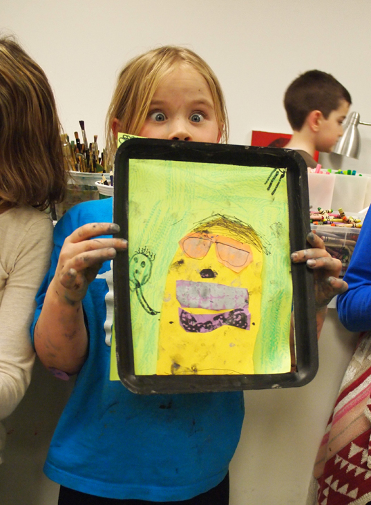 A young girl holds a colourful portrait drawing up over the lower half of her face while her eyes opened comically wide