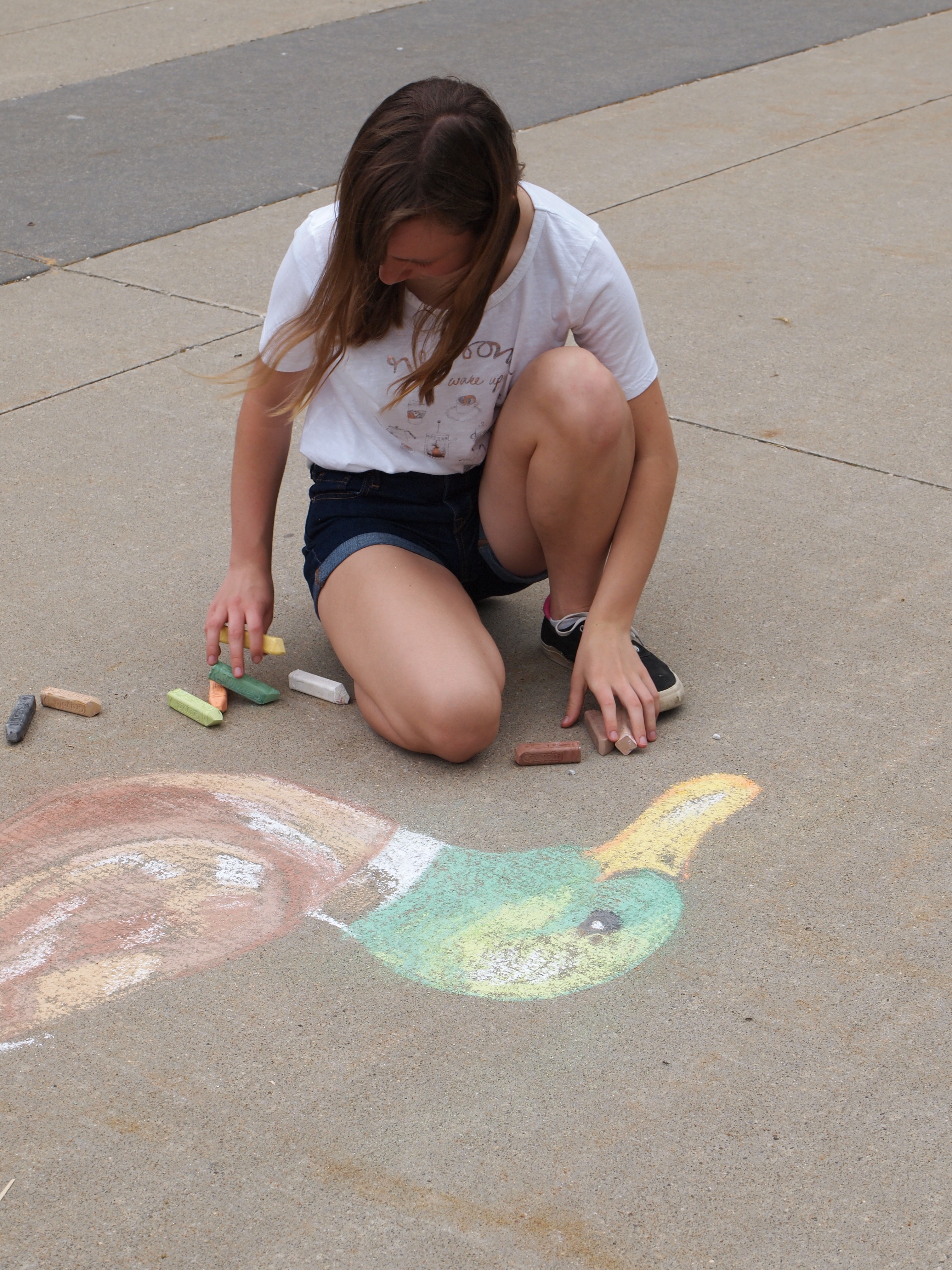 A young girl kneels on concrete alongside a chalk drawing of a duck; pieces of chalk sit on the ground close at hand