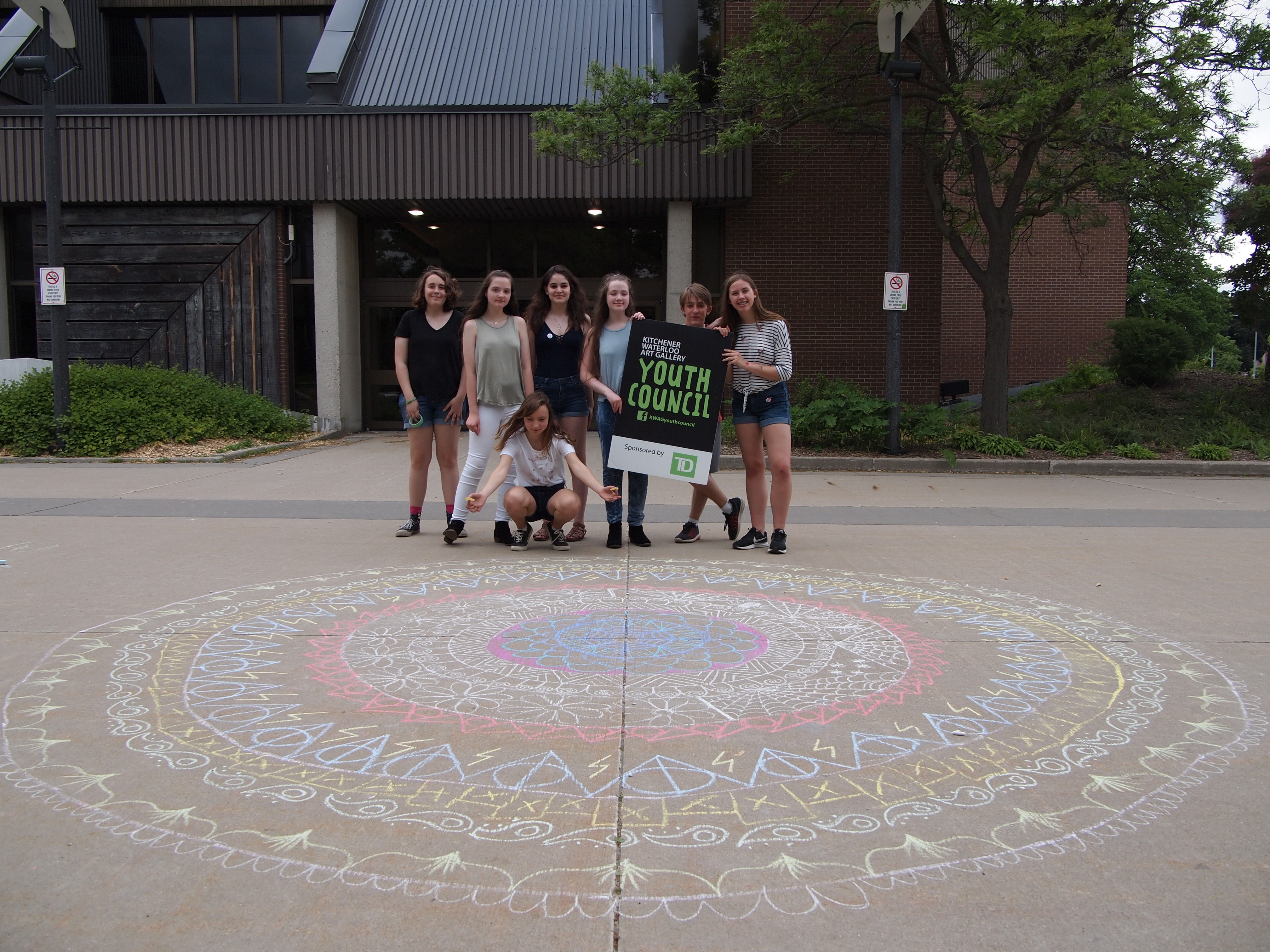 A group of seven youth hold a large sign for the KWAG Youth Council from behind a large chalk mandala drawn on the forecourt outside Centre In The Square