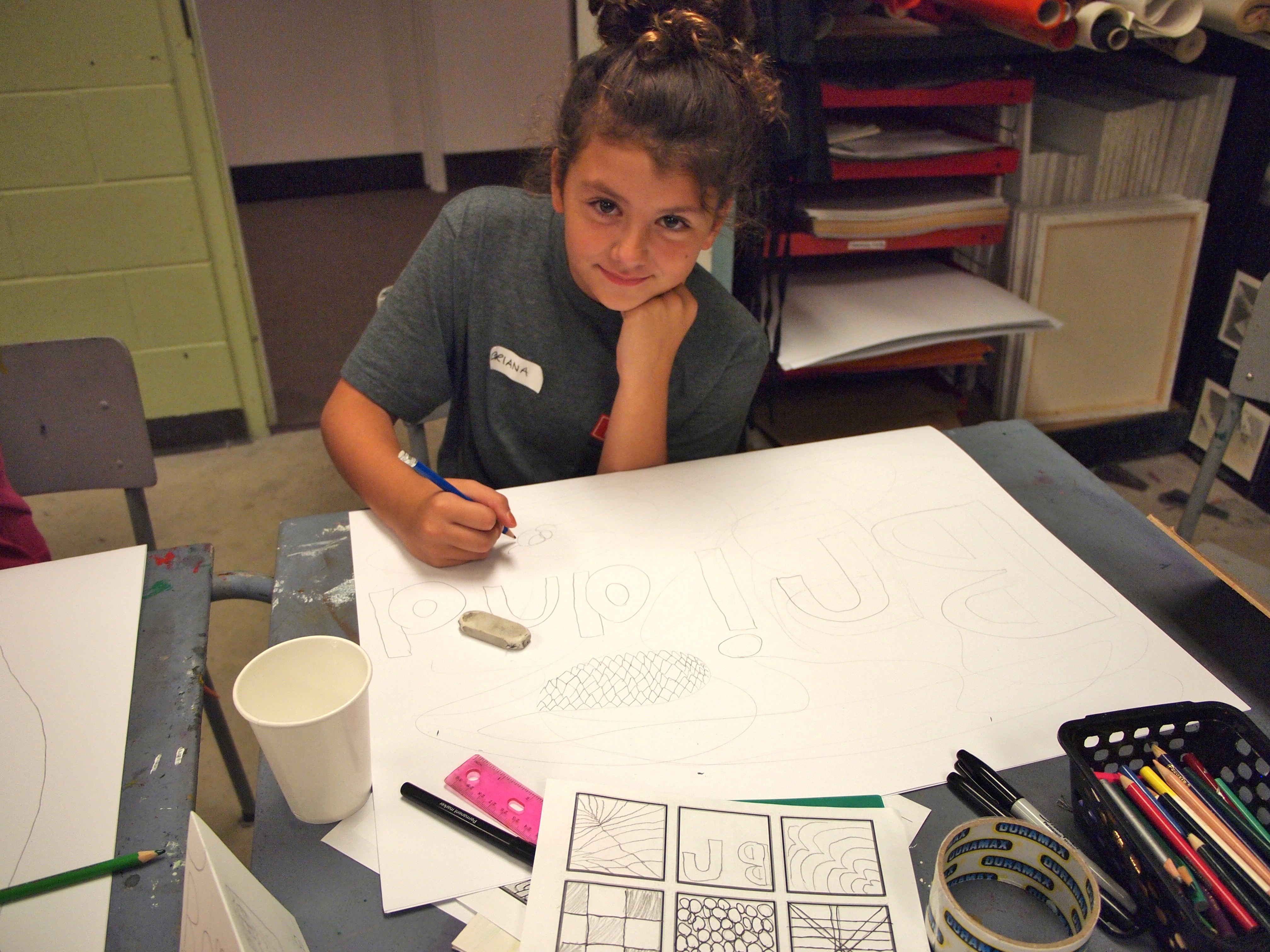 A young girl looks up at the camera as she works on a preliminary drawing on a large sheet of white paper with storyboards laid out in the foreground as reference