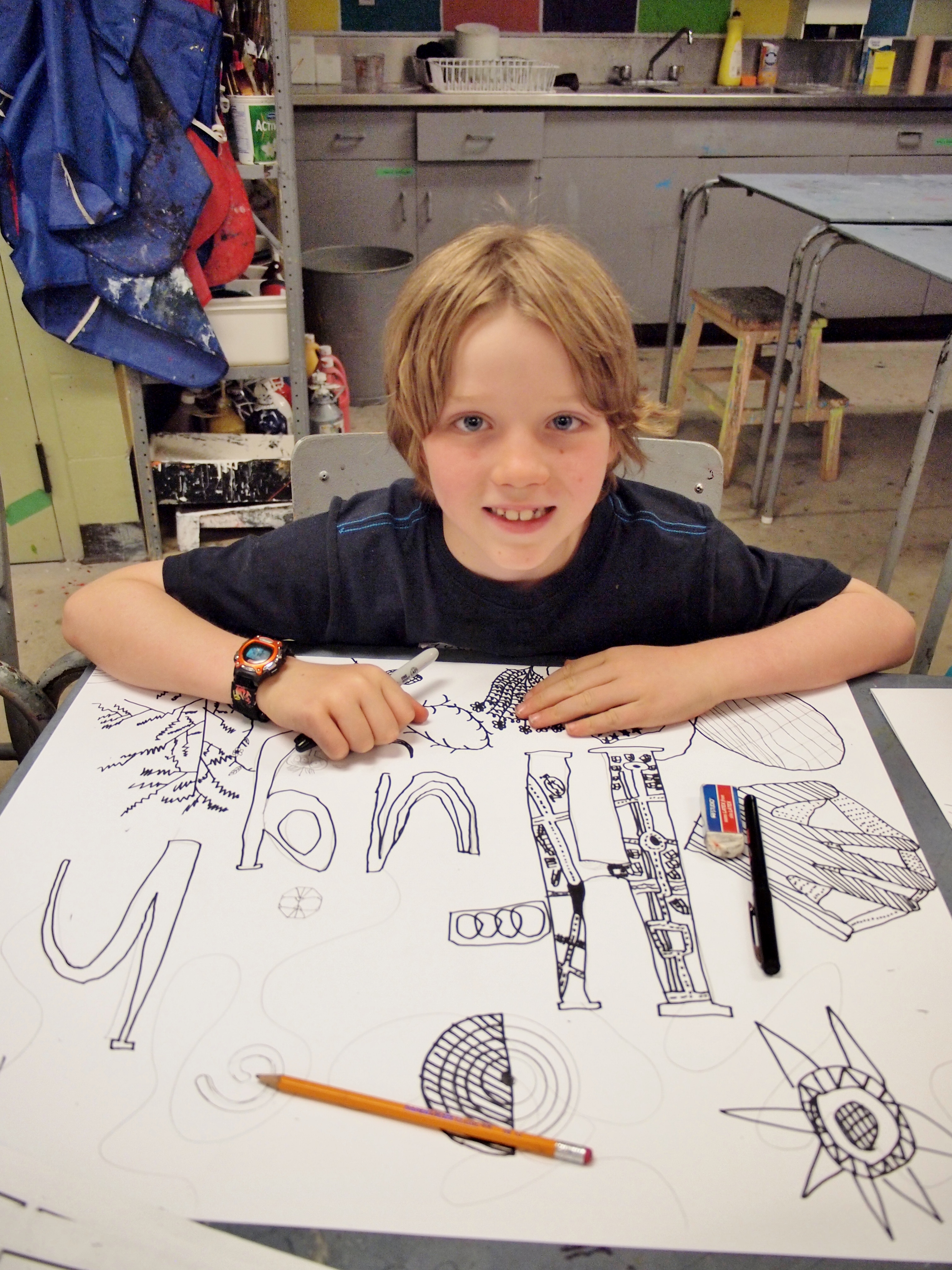 A young child with short blond hair sits at a table looking up from a large black marker drawing of intricate letter shapes