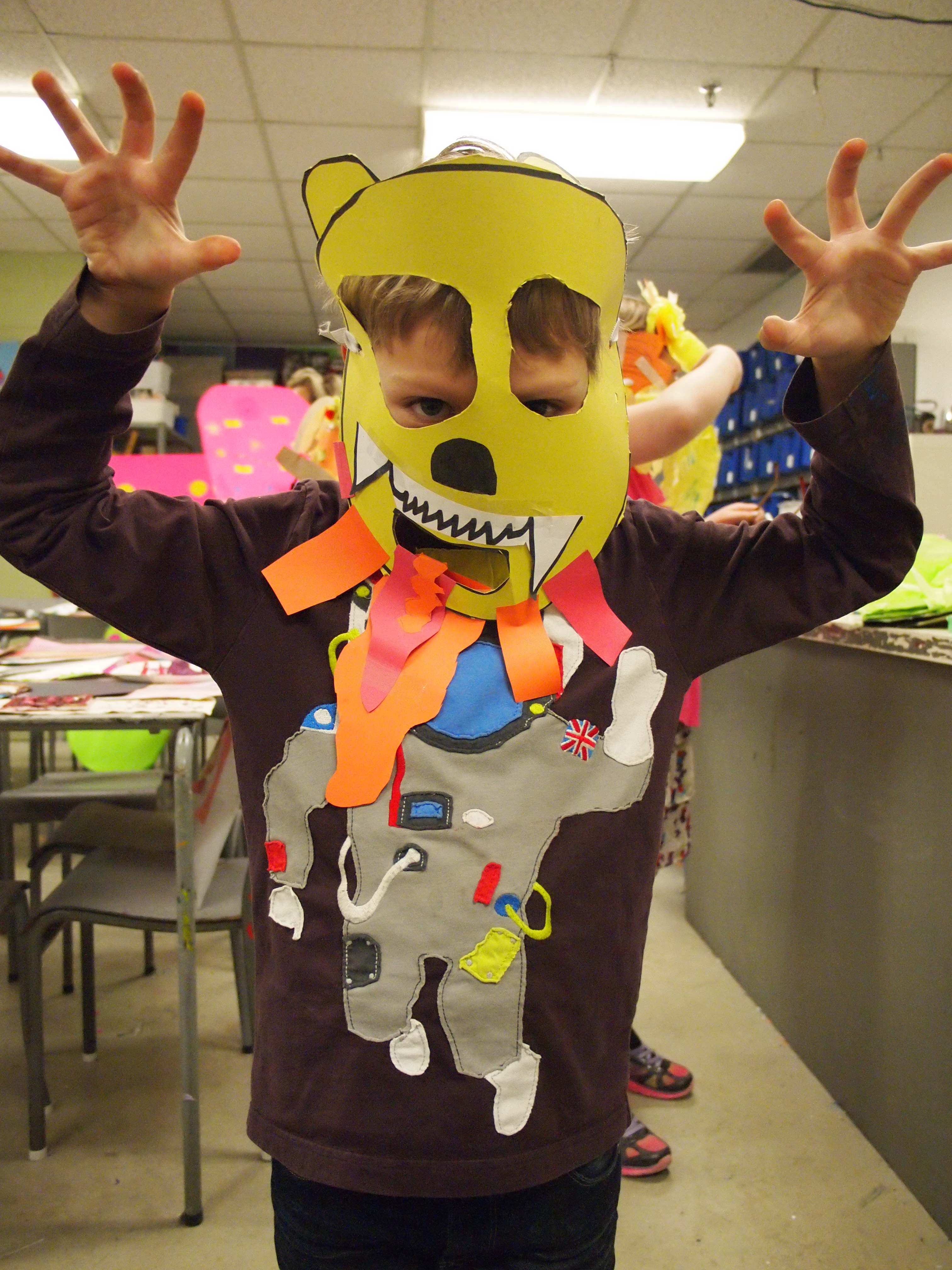A young boy wearing a yellow paper monster mask poses with hands raised like claws