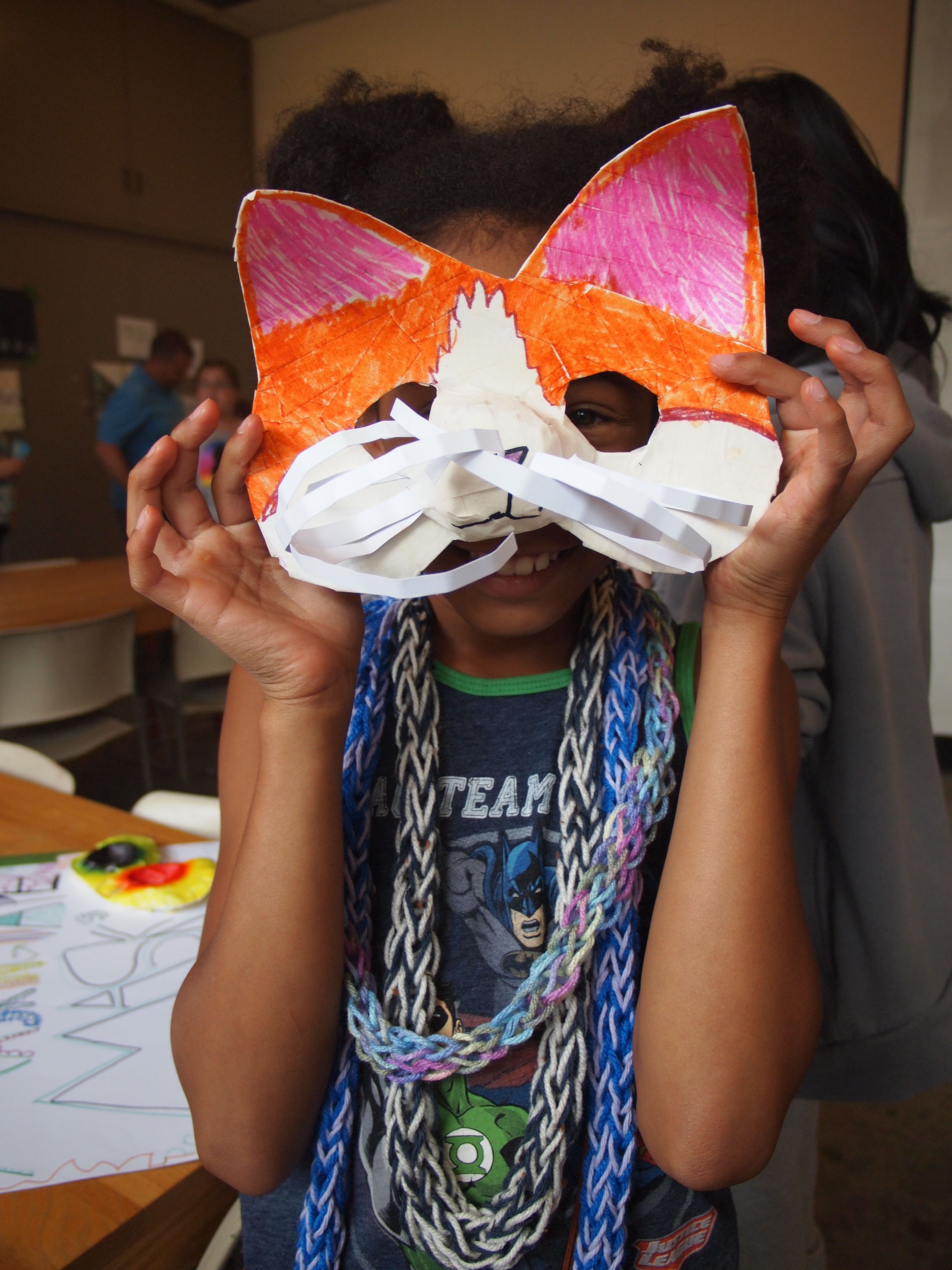 A young girl wearing multiple woven yarn necklaces holds an orange and white cat mask over her face
