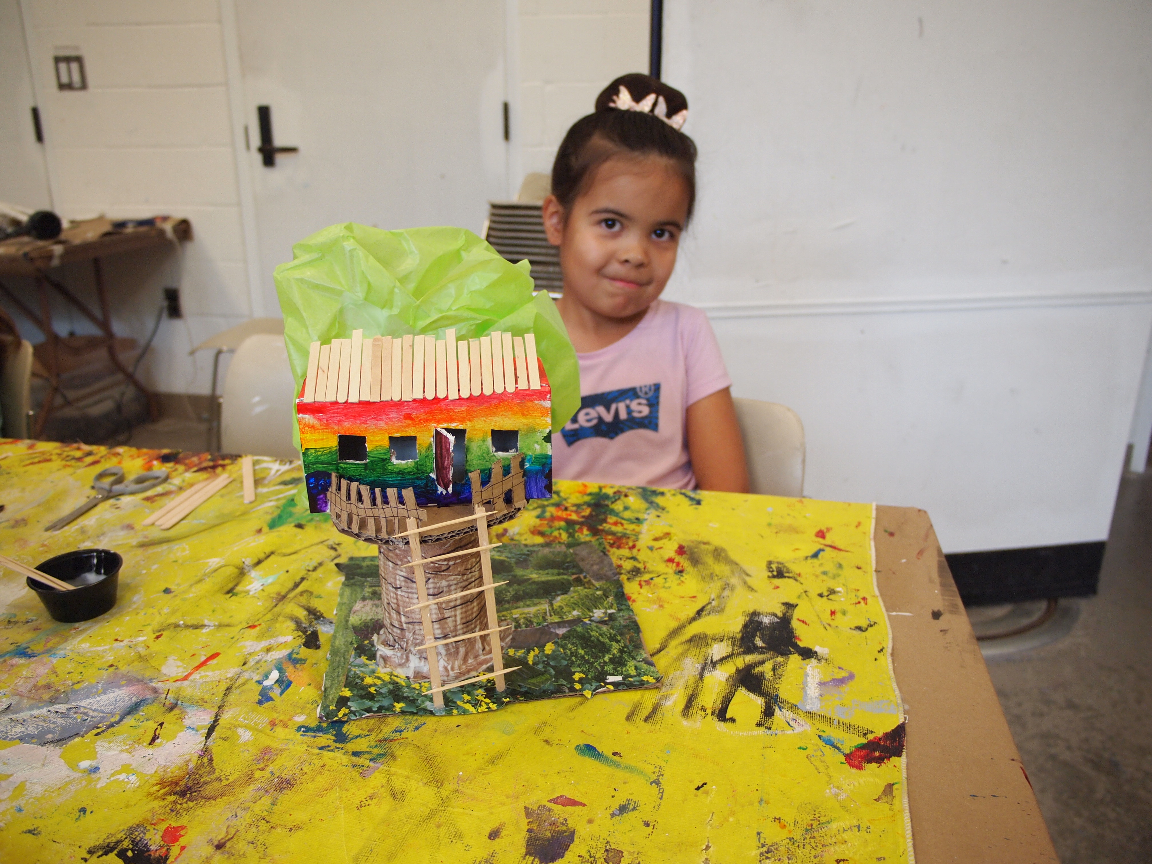 A young girl in a pink Levi's t-shirt smiles while seated behind a mixed-media treehouse sculpture resting on a paint-covered yellow tablecloth