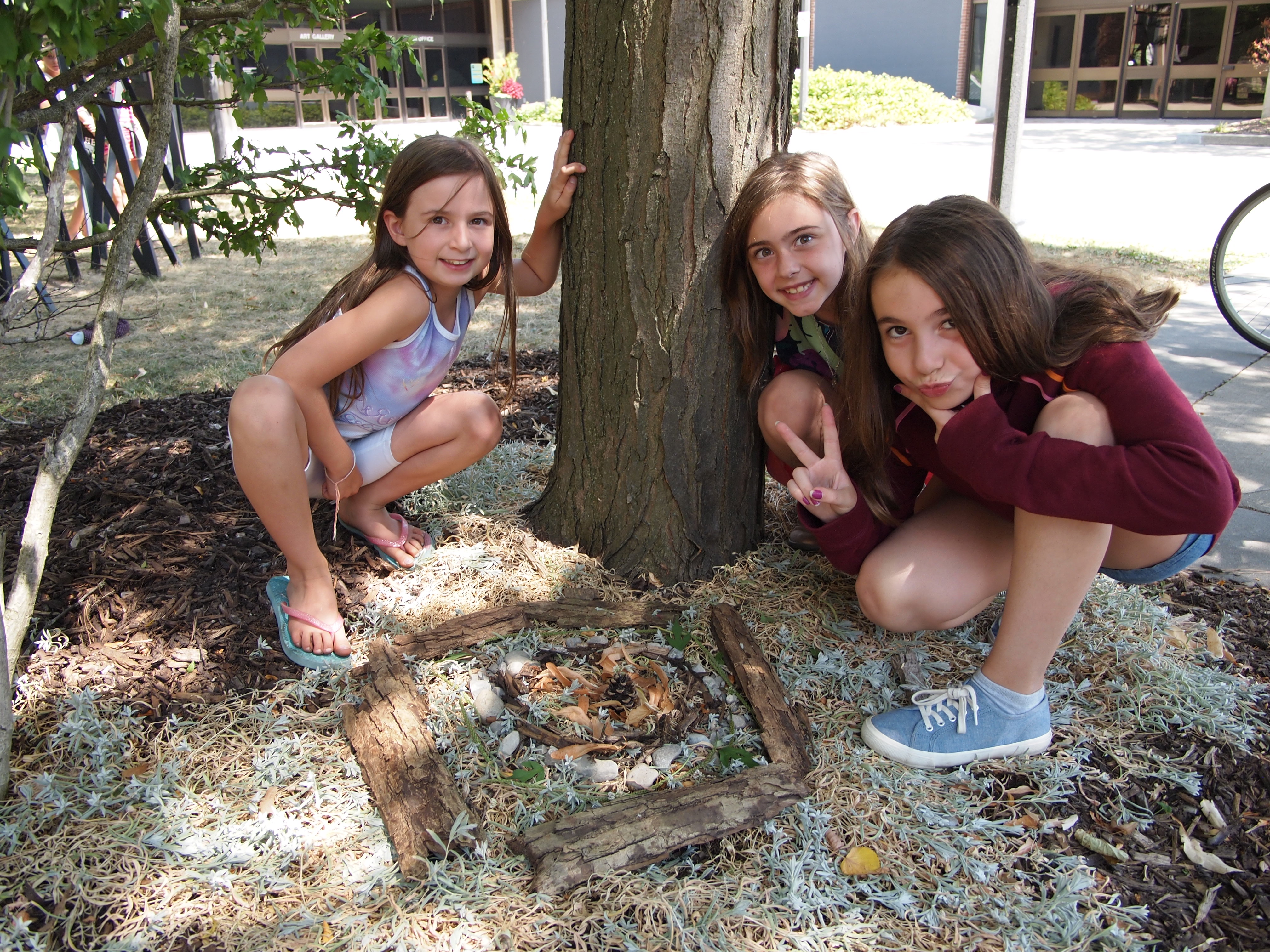Photo of three young girls crouched around the base of a tree smiling at the camera with an artful arrangement of natural materials displayed between them on the ground