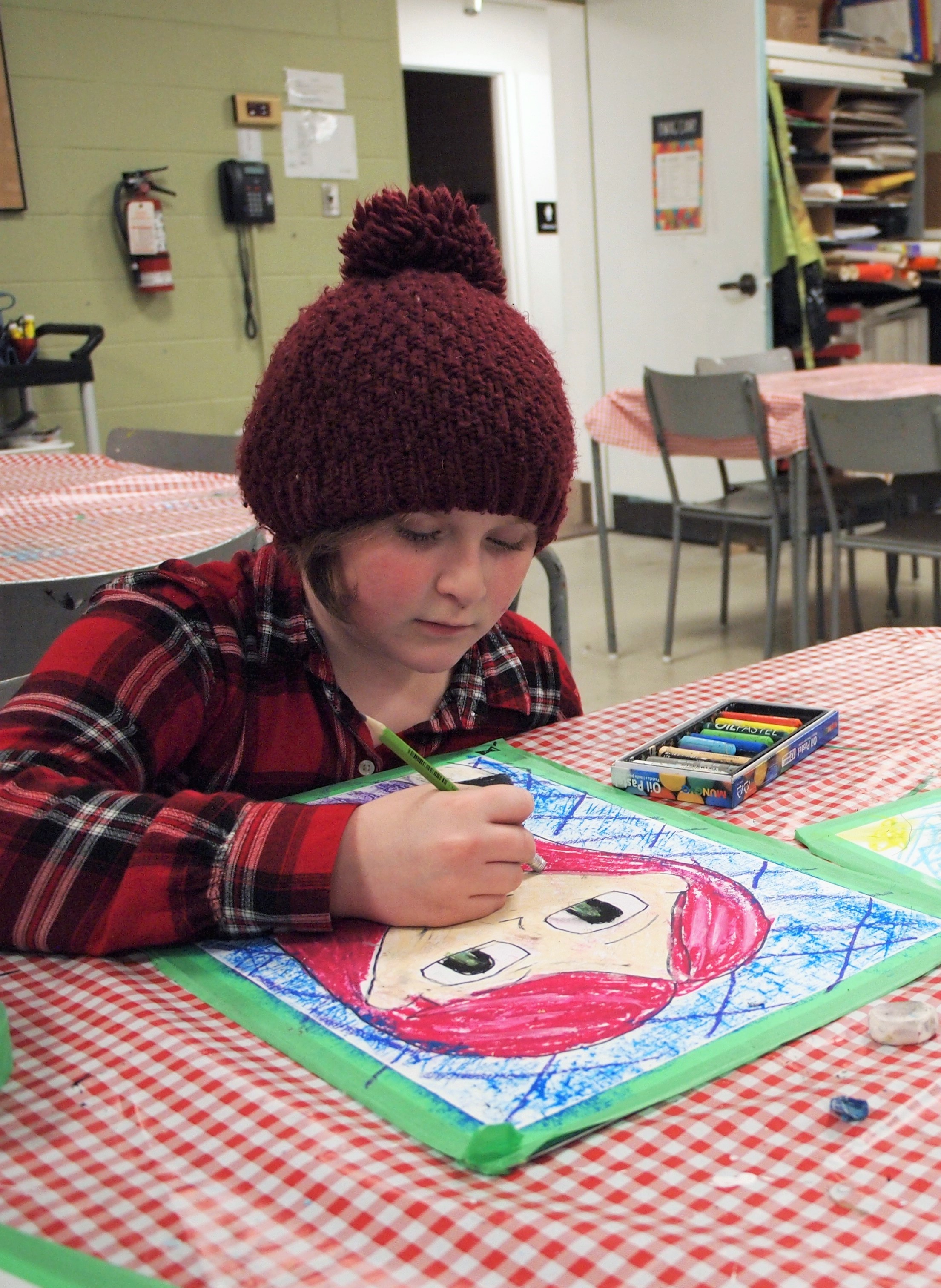 A young girl in a plaid shirt and wooly hat concentrates on drawing a colourful self-portrait