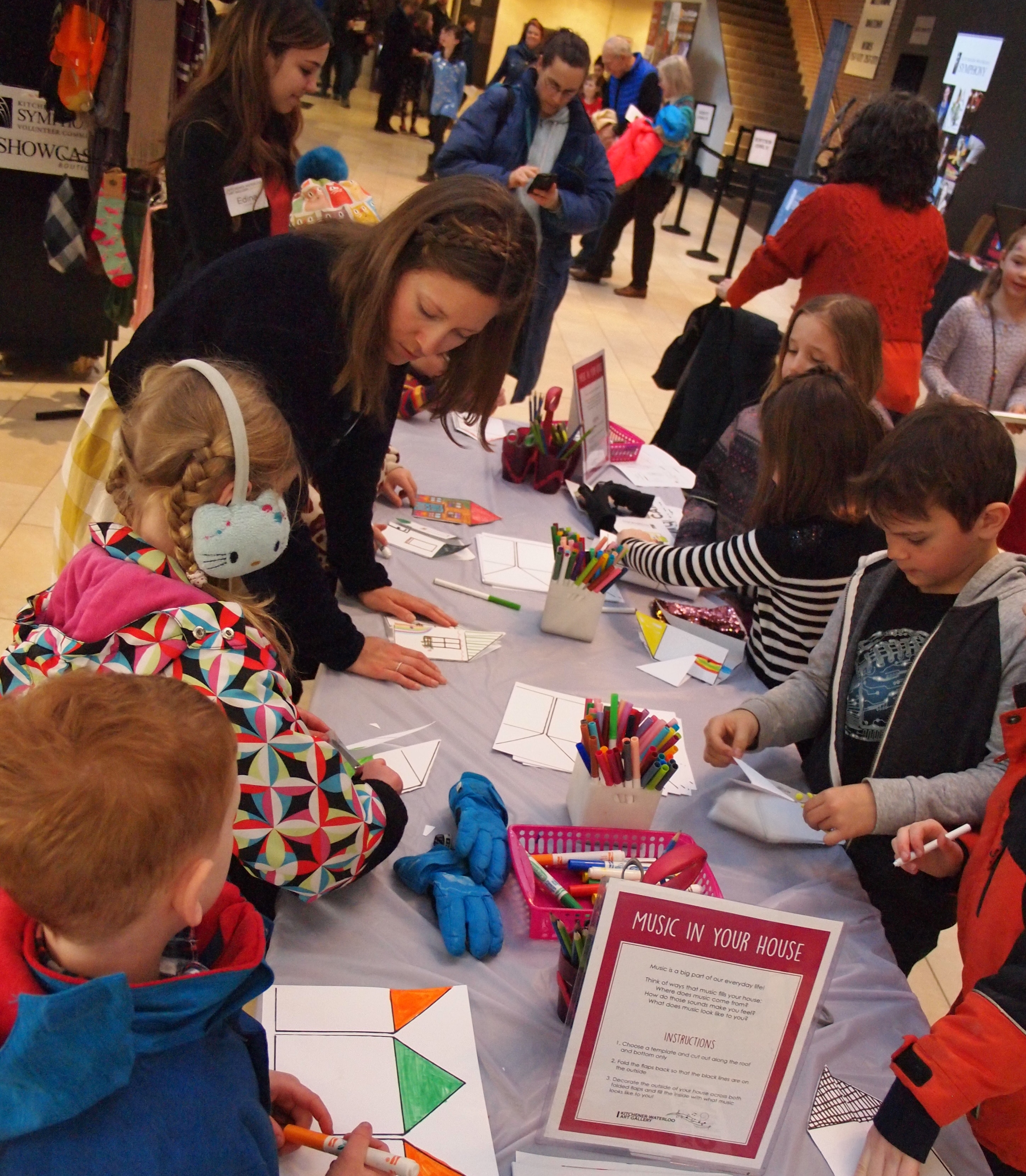 A group of children around a busy craft table in the lobby of Centre In The Square working on an art activity with support from a young woman volunteer