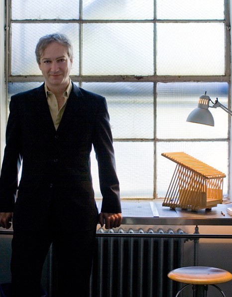 Photo of Paul Raff, an unusually pretty older man in a dark suit and open-collar beige shirt leaning against a windowsill in an industrial building; a small architectural model sits on the sill next to him