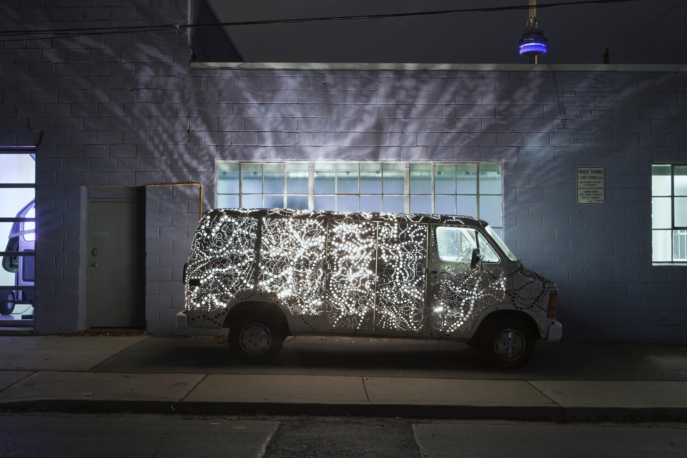 Kim Adams' Auto Lamp is an intricately perforated van displayed on a dark sidewalk, casting ribbons of light on the brick building behind it