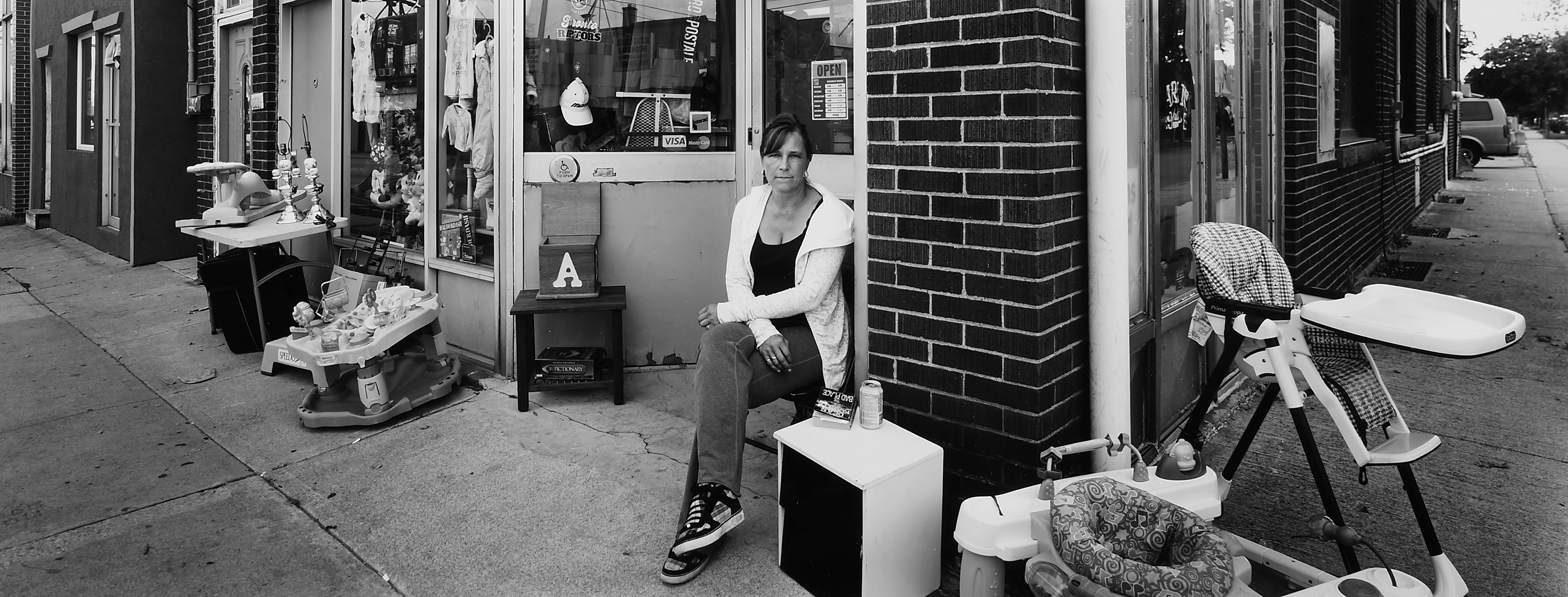 A wide-format black and white photograph centred on a woman seated in storefront cluttered with merchandise both on the sidewalk and in the window