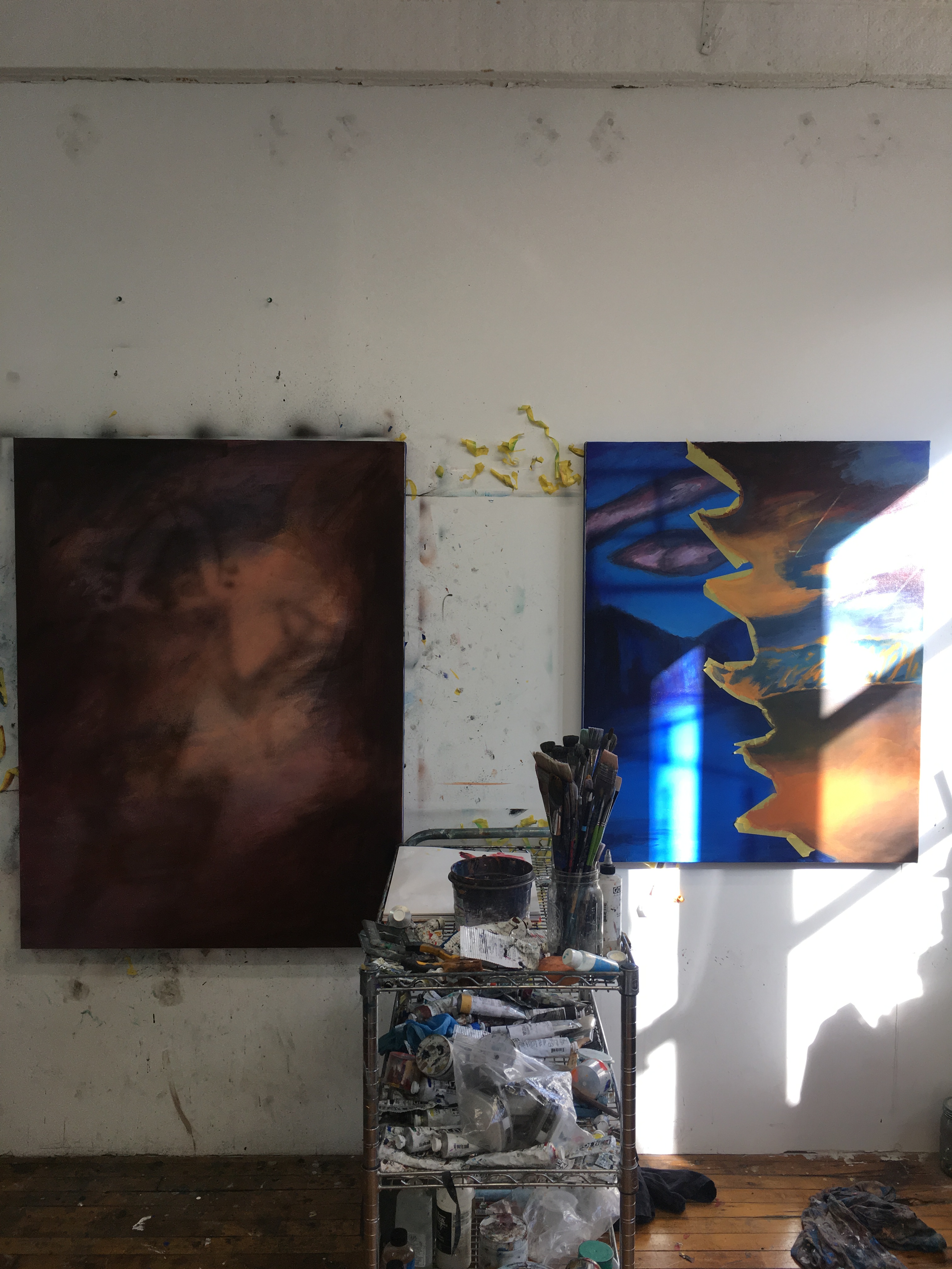 A view of geetha thurairajah's studio showing two paintings in progress hung on a wall, with a messy paint cart standing between them, partly illuminated by strong daylight slanting from the right