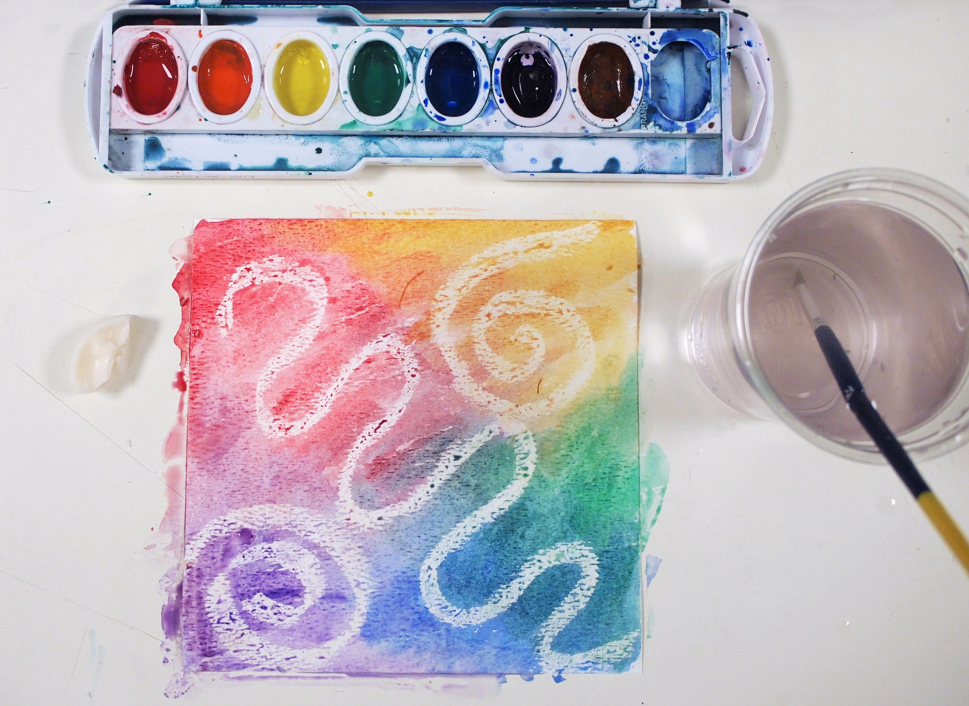 Overhead photo of a colourful watercolour abstract alongside a cup of rinse water and a watercolour palette