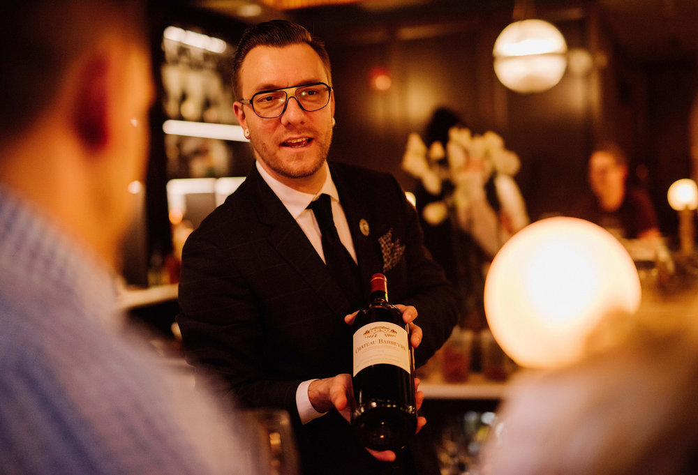 Photo of Wes Klassen, a well-dressed man in contemporary glasses presenting a bottle of wine in a warmly-lit restaurant setting