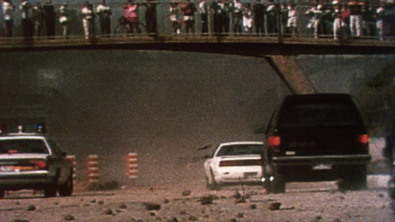 Film still from Rocks at Whiskey Trench showing cars traversing a rock-covered road beneath an overpass lined with protesters