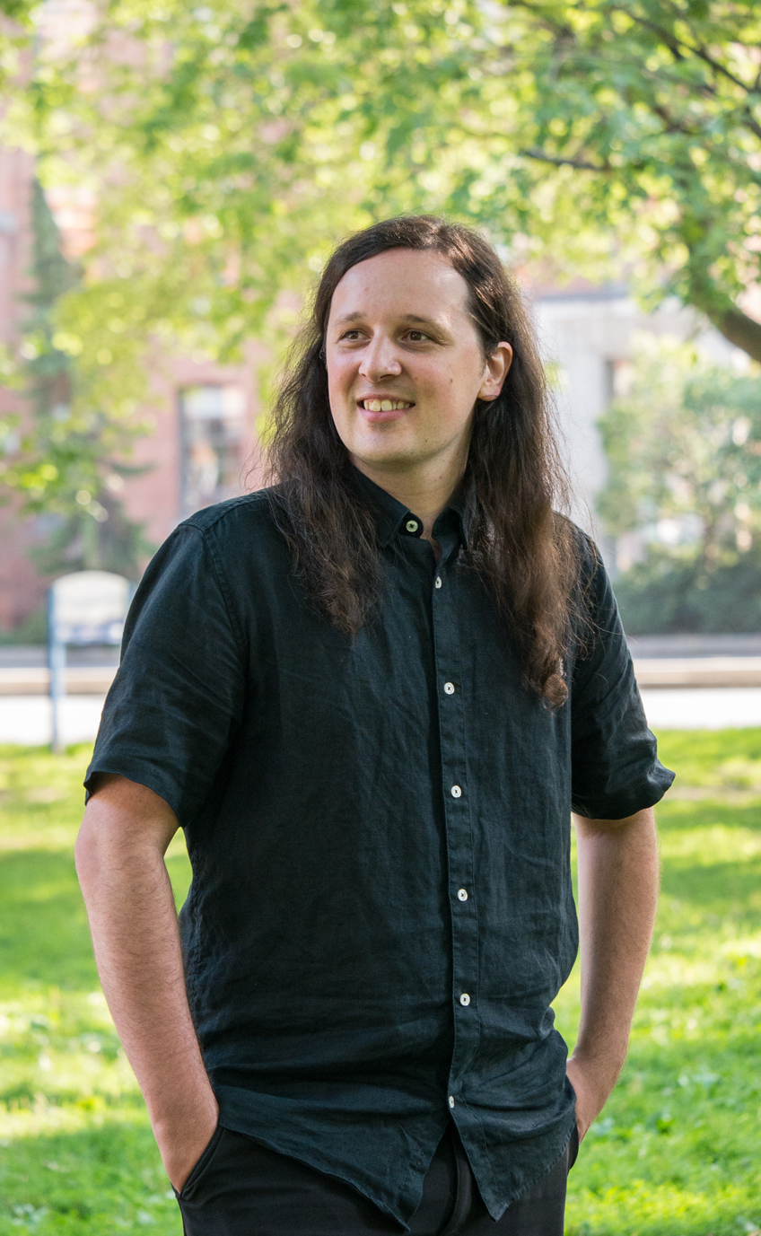 Photo of Darryn Doull, a white man with long, loose brown hair standing outdoors in a public park, wearing a dark short-sleeved shirt and standing with hands in pockets