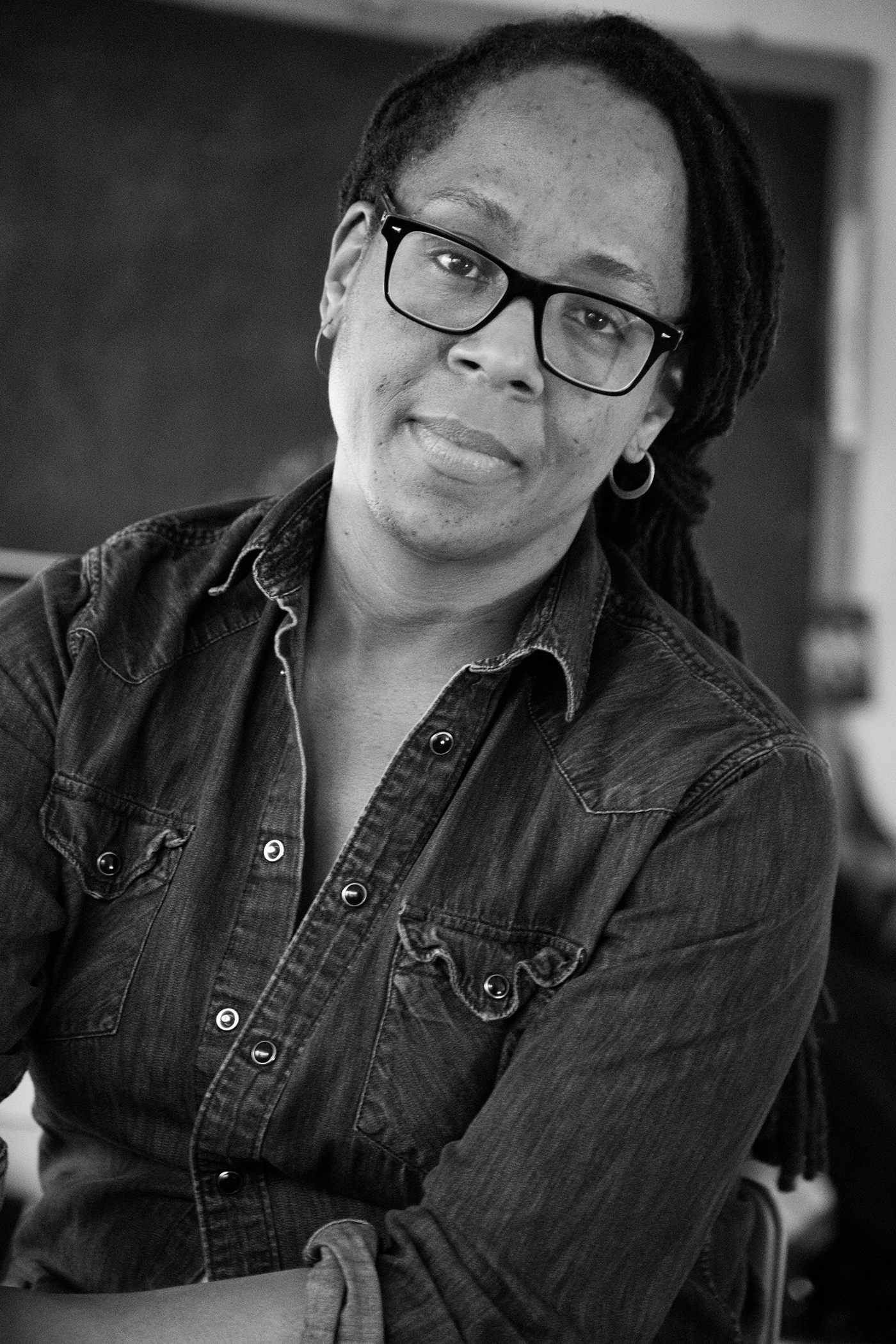 Black and white photo of Deanna Bowen, a Black woman with dark-rimmed glasses, small hoop earrings and long black hair pulled back in braids wearing a button-down denim shirt