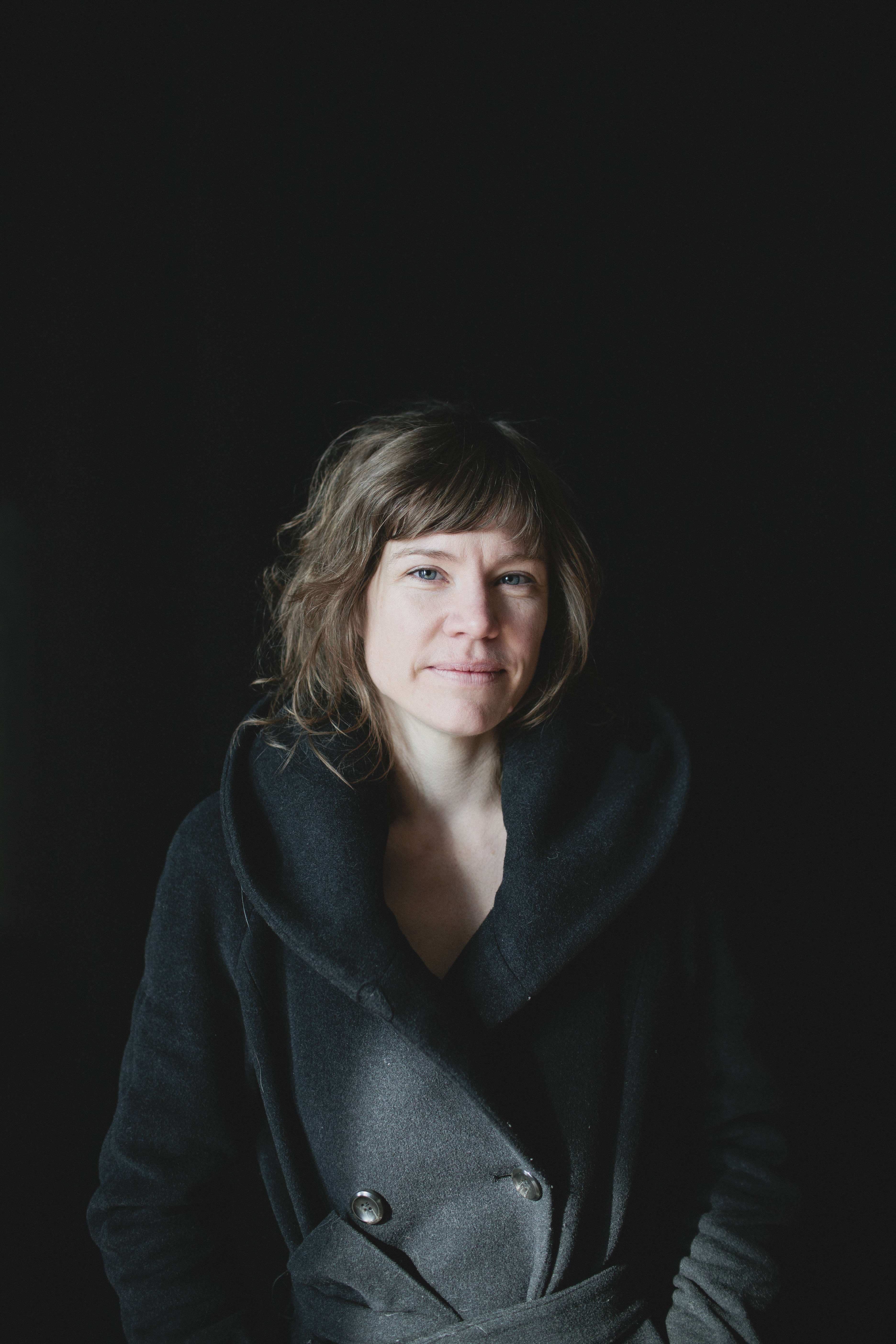 Photo of Katie Ewald, a white woman with light brown hair wearing a heavy black coat with a wide colour, standing against a black background