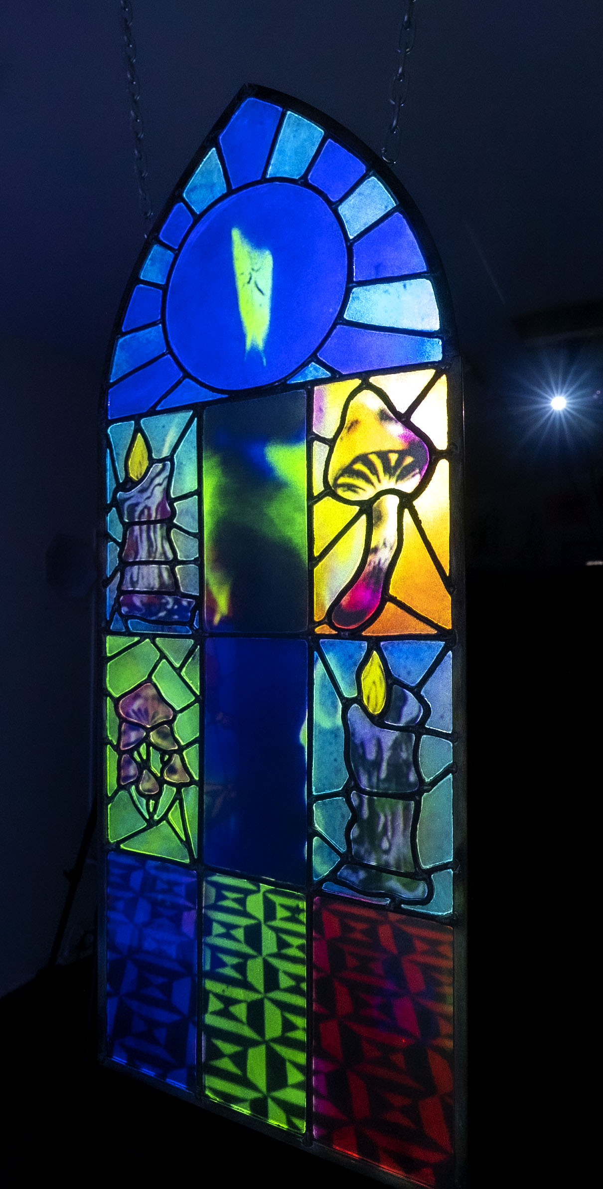 An arched stained glass window illuminated with images of candles, mushrooms and modern tile patterns