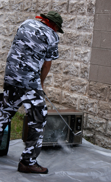 A man in grey camo fatigues smashes a CRT monitor with a golf club