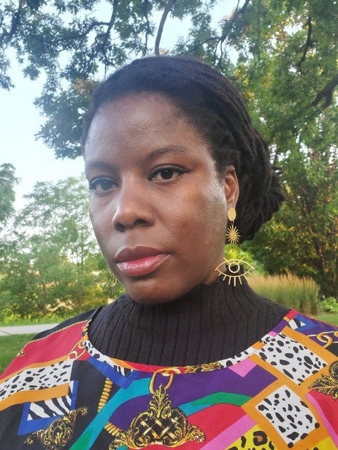 Photo of Glodeane Brown, a Black woman seen standing beneath a tall tree canopy, wearing a brightly patterned top and a large gold wire earring shaped like an eye
