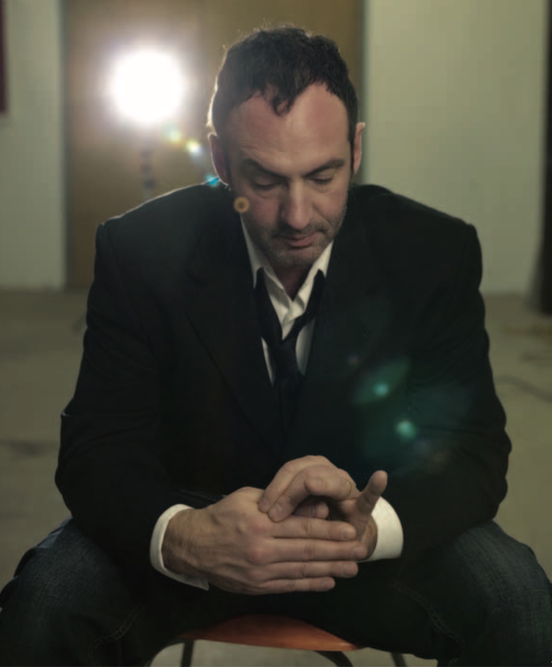 Photo of Milutin Gubash, a dark-haired balding man wearing a suit, seated with head bowed in a space with a strong light source behind him