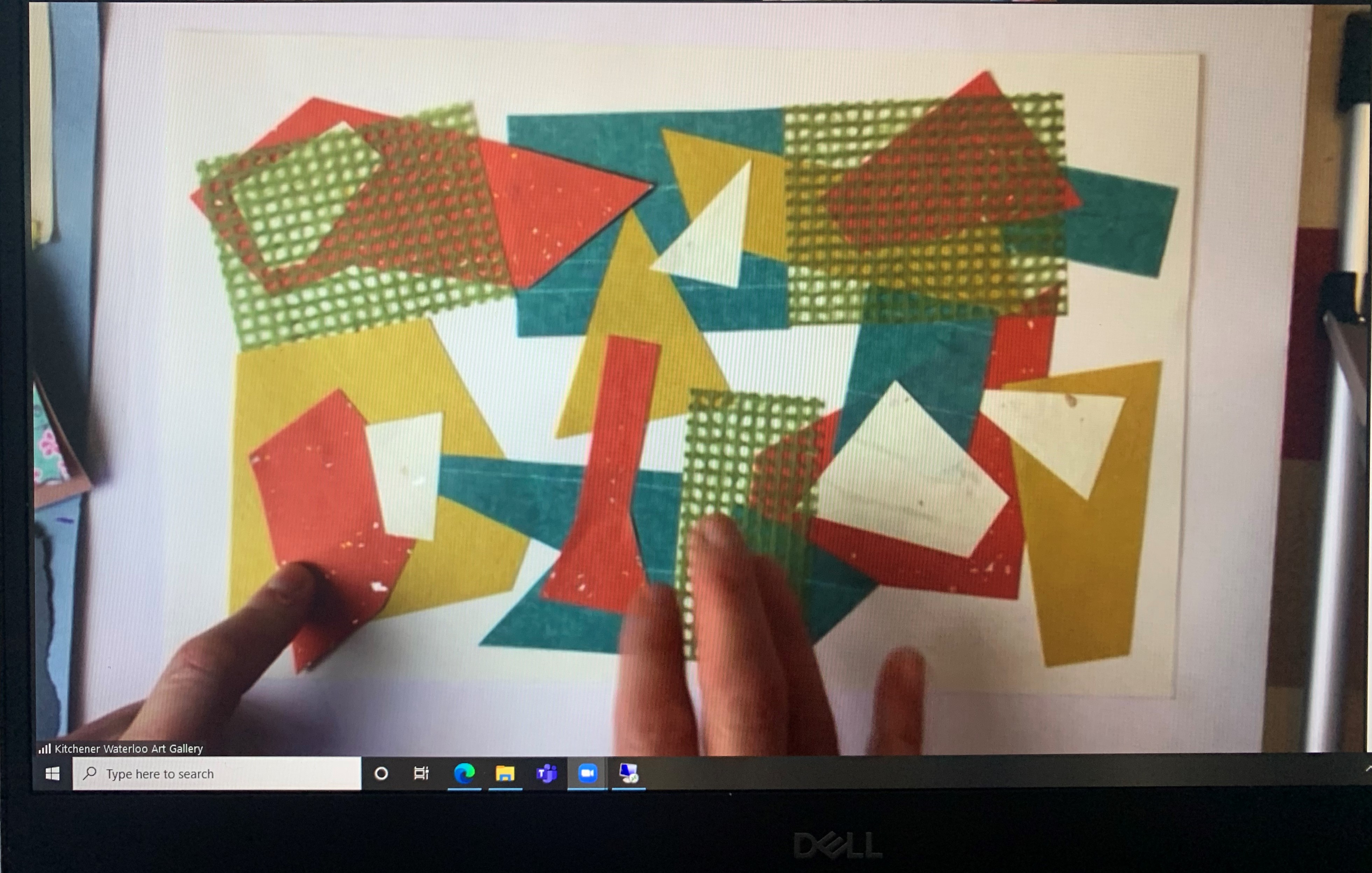 View of a colourful collage in progress with two hands manipulating shapes as seen via a Zoom videoconferencing screen