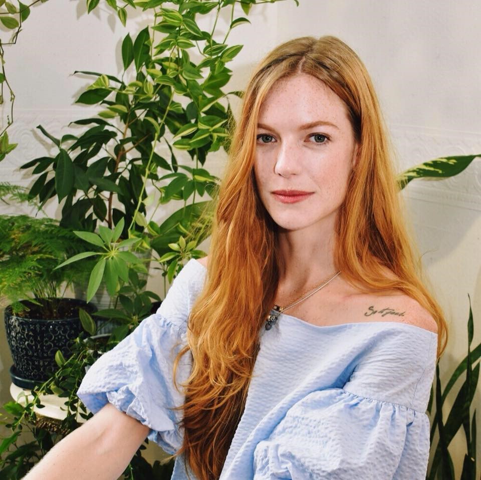 Photo of Angel Callander, women with long red hair, fair complexion wearing a short sleeve top. Houseplants feature in the background.