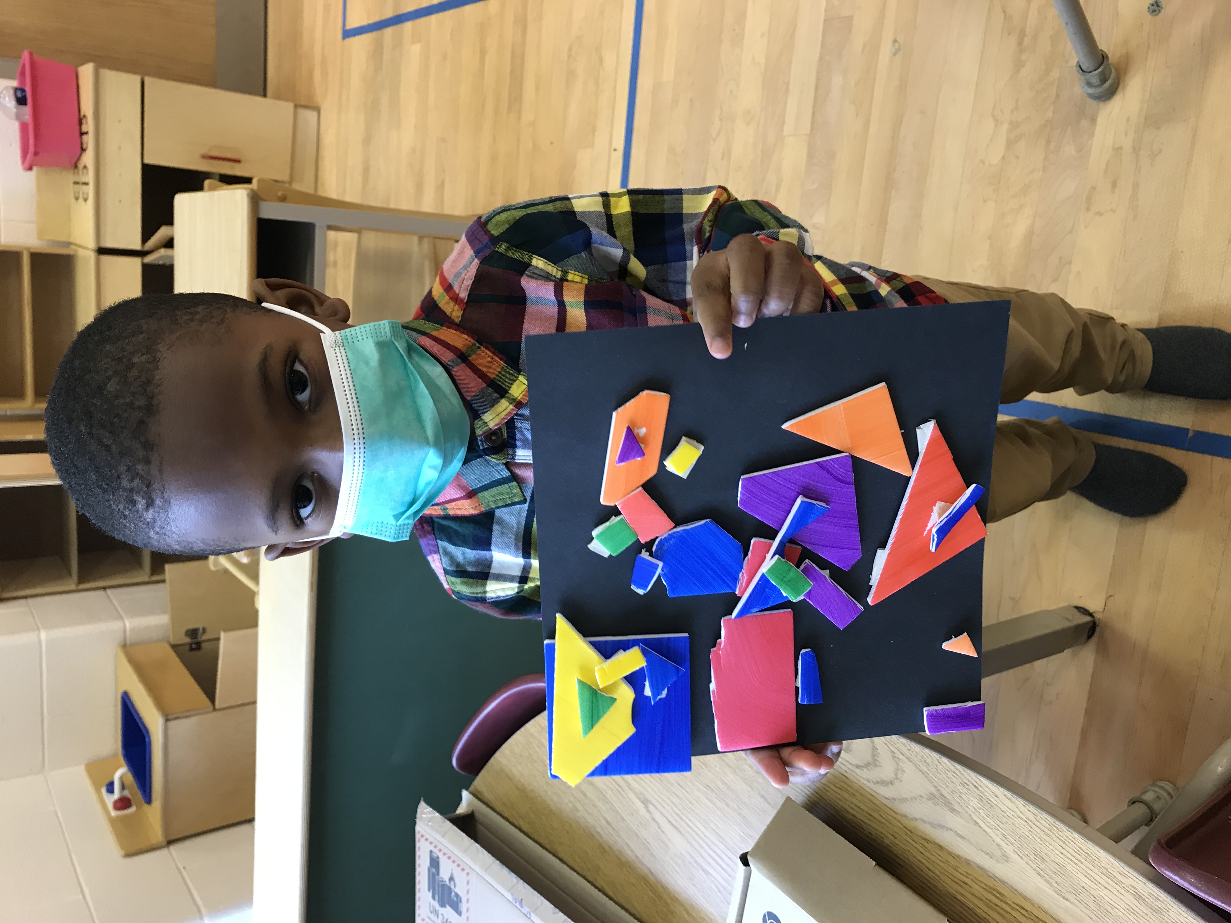 Young boy wearing a face mask holding up a black sheet of paper with colorful shapes layered on top of each other