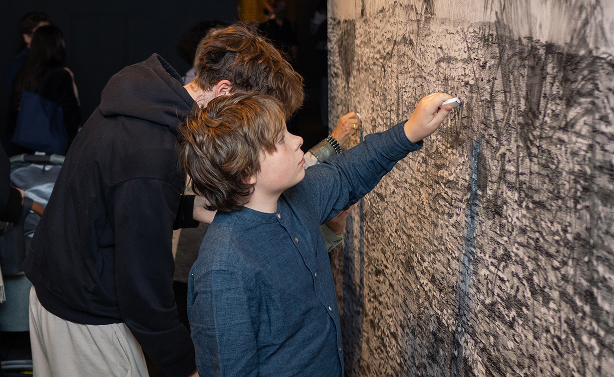 Photo of two male presenting youths using erasers to make a mark on a graphite based artwork by Claire Greenshaw