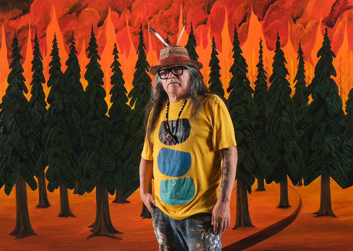 Photo of the artist Lawrence Paul Yuxweluptun standing in front of an abstract painting of a forest in flames