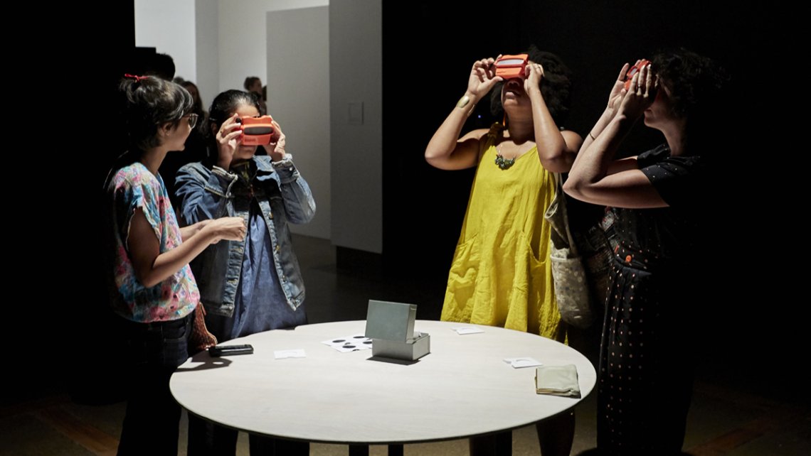 Four women gathered around a round white table in a dark gallery space, looking through slides using Viewmaster toys