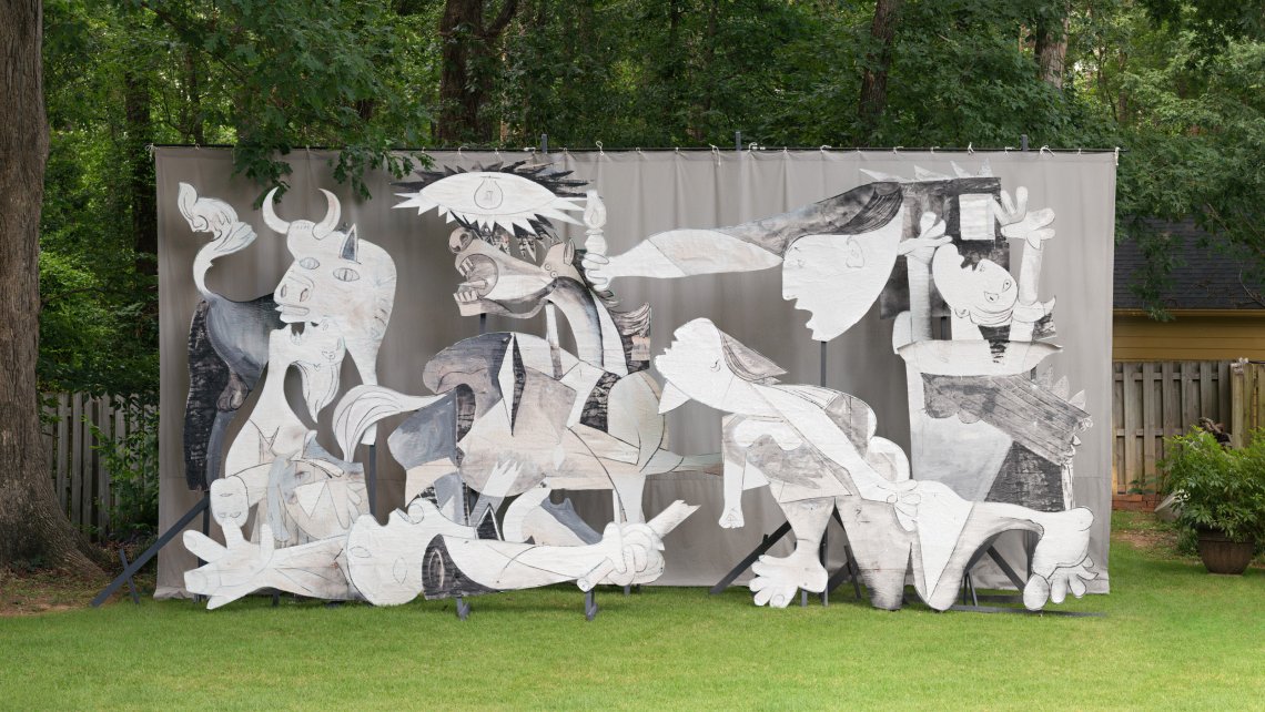 Adad Hannah photo of a restaging of Picasso's Guernica using wood and cardboard cut-outs, arranged in a lush green backyard space