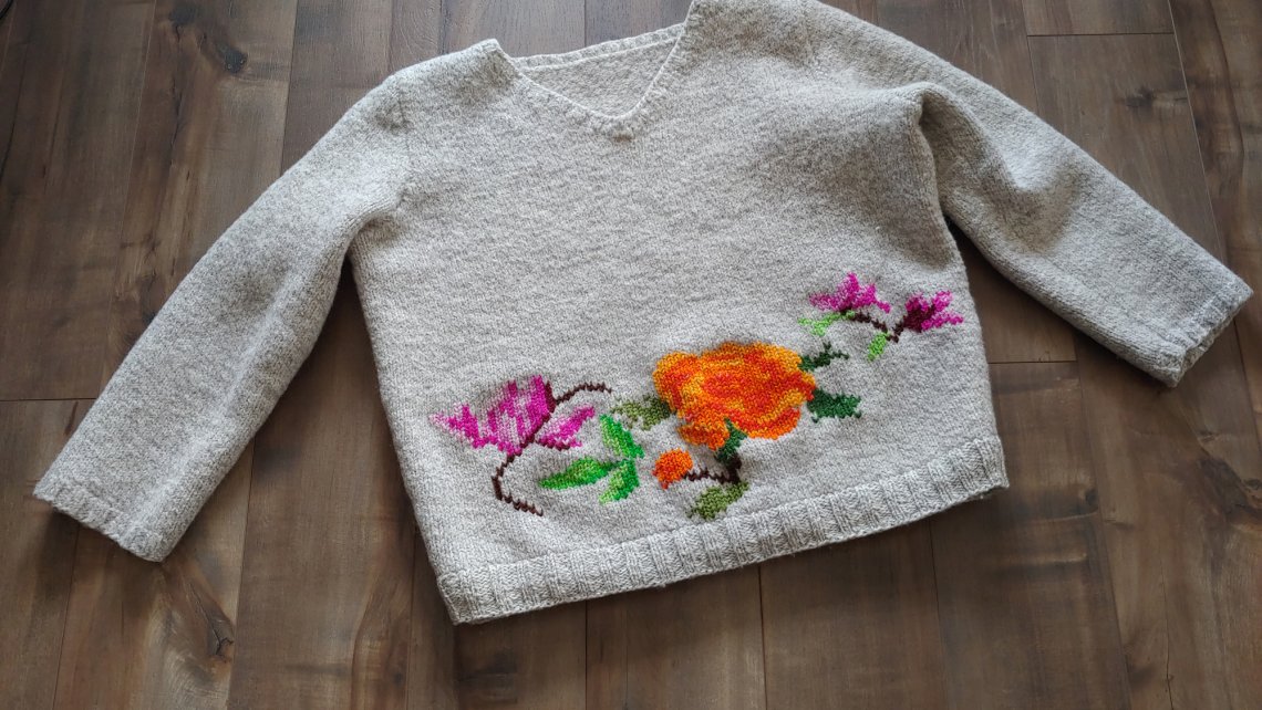 A white knitted sweater spread out on a dark wood laminate floor with a pattern of violet and orange flowers worked into its lower edge