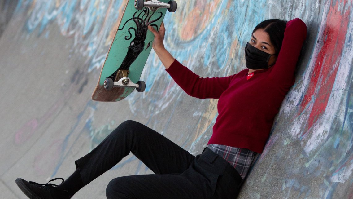 A female skateboarder in dark clothes and a black facemask holds up her green skateboard while reclined against the side of a graffiti-covered half-pipe wall