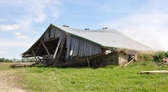 A photograph of a collapsing wood barn in a bright spring landscape of green grass and blue sky