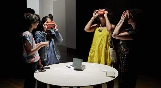 Four women gathered around a round white table in a dark gallery space, looking through slides using Viewmaster toys