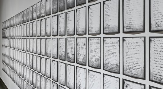 A sidelong view of Deanna Bowen's prints of a 1911 petition, showing black-edged letter-sized pages receding in rows along a white gallery wall