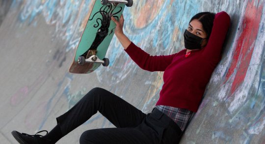 A female skateboarder in dark clothes and a black facemask holds up her green skateboard while reclined against the side of a graffiti-covered half-pipe wall
