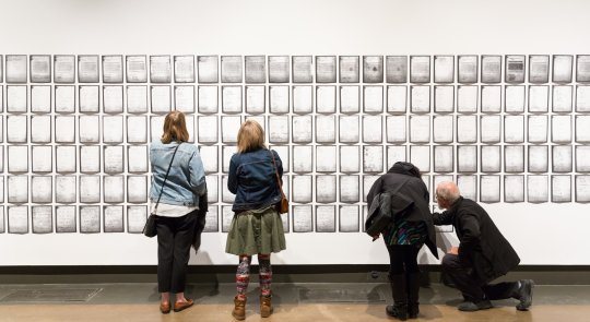 Photo of two pairs of gallery visitors seen from behind as they examine rows of pages of Deanna Bowen's installation of a 1911 petition, which fills the wall to both edges of the photo