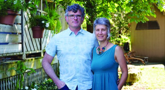 Photo of Trent Bauman and Juanita Metzger, an older couple casually dressed and standing together in a backyard filled with lush greenery