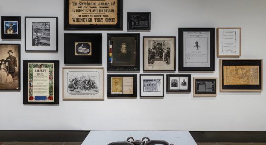 Installation view of Deanna Bowen's Black Drones in the Hive, showing a pair of shackles displayed on a white plinth in front of a wall filled with framed archival images