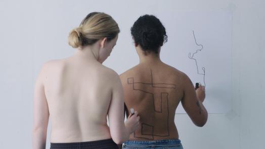 Two women seen from behind, nude from the waist up, as one draws lines on the other's back that the recipient translates to lines drawn on the white wall before them