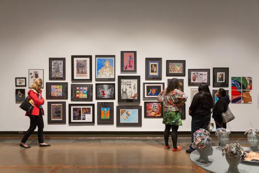 Gallery visitors pause in front of a large wall featuring a salon-style hanging of over 20 framed student artworks; a low table displaying sculptures is visible in the lower right foreground