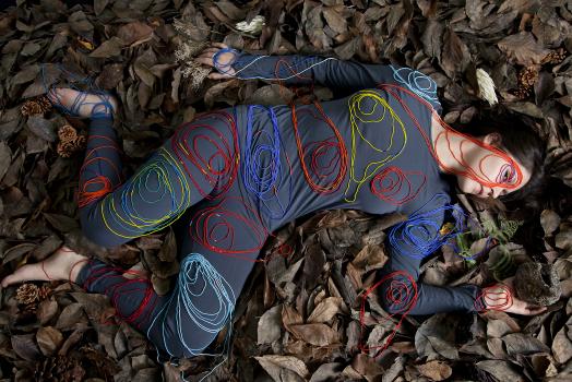 Meryl McMaster's Terra Cognitum depicts the artist in a dark blue leotard, reclining in a bed of winter leaves with circular motifs drawn in coloured thread over her body; her head is turned to the side while her eye returns the viewer's gaze