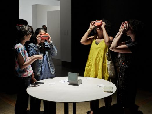 Four women gathered around a round white table in a dark gallery space, looking through slides using Viewmaster toys